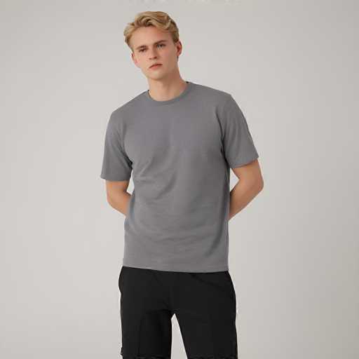 A fair-skinned man with short, light blonde hair is centrally positioned against a plain white background. He's wearing a heather gray t-shirt and black pants, with his hands clasped behind his back. The lighting is soft and even, creating a clean, minimalist aesthetic typical of fashion product photography.