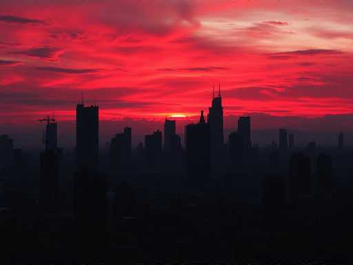 A breathtaking cityscape at sunset, where the sky is ablaze with a mesmerizing palette of reds, pinks, and purples. The sun, partially obscured by clouds, casts a warm glow over the scene, creating a stunning contrast against the dark silhouettes of the towering skyscrapers below. The interplay of light and shadow highlights the architectural grandeur of the buildings, while the vibrant sky adds a dramatic and ethereal quality to the urban landscape.