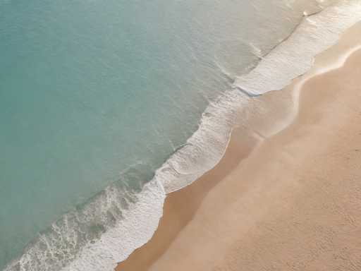 This aerial photograph captures a serene beach scene, showcasing the interplay between the ocean and the sandy shore. The image is divided diagonally from the bottom left to the top right, with the ocean occupying the upper left portion and the sandy beach in the lower right. The ocean water is a light blue, almost turquoise, with gentle waves creating white foam as they lap against the shore. The sand is a light tan color, appearing wet and smooth, with subtle ripples and footprints visible. The sunlight, coming from the top right, casts a warm glow over the entire scene, enhancing the tranquil and picturesque quality of the beach.
