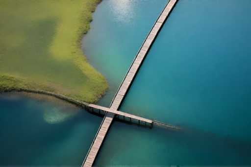 The image shows an aerial view of a wooden bridge crossing over a body of water. The bridge is made up of multiple planks and spans the width of the river, connecting two points on either side. The surrounding area appears to be lush with vegetation, including grass and trees that line both sides of the river.