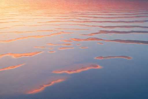 This image captures a mesmerizing aerial view of a vast body of water, likely an ocean or a large lake, during either sunrise or sunset. The water's surface is adorned with gentle ripples and waves, creating a dynamic interplay of light and shadow. The sky is painted in a stunning palette of colors, with the upper left corner bathed in a warm yellow glow, transitioning to a rich orange hue, and finally blending into a deep purple as it stretches across the horizon. The ripples on the water's surface reflect these vibrant colors, adding a shimmering effect to the scene. The overall composition is serene and captivating, showcasing the natural beauty of the moment.