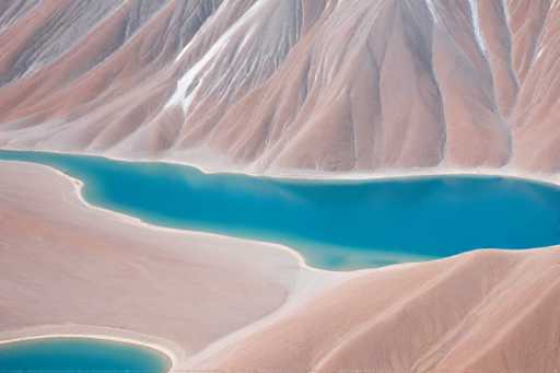 The image shows an aerial view of a mountain range with two lakes nestled within the mountains. The lake on the left is a light blue color and appears calm, while the lake on the right has a darker blue hue and seems to be more active. The mountains surrounding the lakes are covered in brown and white rock formations that vary in size and shape.