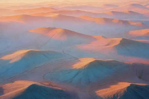 This image captures a breathtaking aerial view of a vast desert landscape, likely taken from a high vantage point such as an airplane or helicopter. The scene is dominated by undulating sand dunes that stretch endlessly into the horizon, creating a mesmerizing pattern of peaks and valleys. The dunes are bathed in the warm, golden light of a setting sun, which casts a beautiful orange glow across the entire landscape. The interplay of light and shadow accentuates the contours of the dunes, giving them a textured appearance with subtle blue hues in the shadows. The sky above is a clear, vibrant blue, contrasting sharply with the earthy tones below. There are no signs of human presence or vegetation, emphasizing the raw, untouched beauty of this natural environment. The overall effect is a serene and awe-inspiring vista that highlights the grandeur of the desert at dusk.