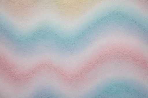 The image depicts a close-up view of a textured surface, possibly resembling a wall or ceiling, characterized by its rough and uneven texture. The surface is adorned with a series of wavy, zigzag lines that create a pattern of alternating colors. Starting from the top, the colors transition from a light brown to a light blue, followed by a white section, then a darker blue, another white section, and finally a pink hue. This pattern repeats with variations, including a darker pink and a lighter blue towards the bottom. The colors are not sharply defined but rather blend into each other, creating a soft gradient effect. The overall appearance is reminiscent of a watercolor painting, with the colors appearing to bleed into one another, enhancing the fluid and dynamic nature of the image.